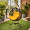 A single hanging rattan swing chair with vibrant yellow cushions on a modern balcony featuring artificial turf and potted palms.