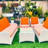 A large white rattan 7-seater conversation sofa set including a sofa, loveseat, two chairs, and a glass table with vibrant orange and geometric patterned cushions on a green lawn.