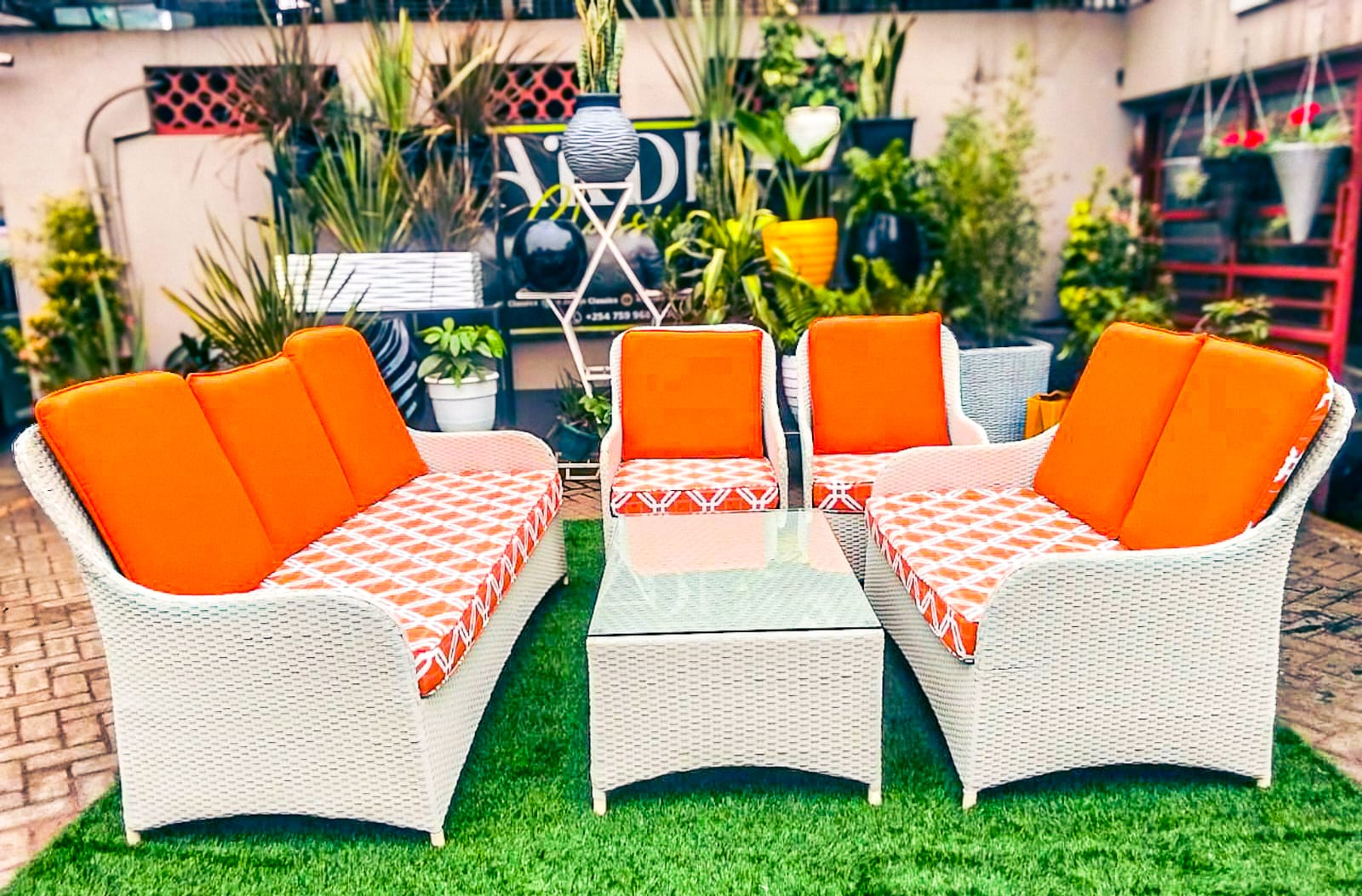 freeesia A large white rattan 7-seater conversation sofa set including a sofa, loveseat, two chairs, and a glass table with vibrant orange and geometric patterned cushions on a green lawn.