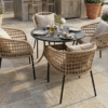 A 4-seater artisan rope weave dining set with a round charcoal table and beige cushions on a luxury stone patio surrounded by Mediterranean plants.