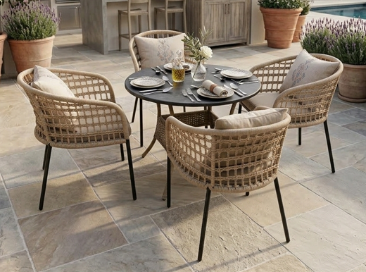 Screenshot 2026-03-23 122122 A 4-seater artisan rope weave dining set with a round charcoal table and beige cushions on a luxury stone patio surrounded by Mediterranean plants.