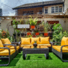 A four-piece outdoor rattan furniture set with bright yellow cushions sits on an artificial turf patch in an urban patio. The set includes a sofa, two armchairs, a love seat and a coffee table. The patio is decorated with many potted plants and a geometric brick wall.
