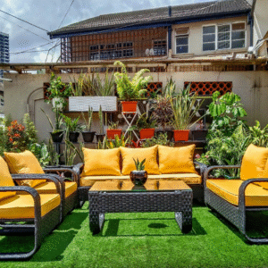 A four-piece outdoor rattan furniture set with bright yellow cushions sits on an artificial turf patch in an urban patio. The set includes a sofa, two armchairs, a love seat and a coffee table. The patio is decorated with many potted plants and a geometric brick wall.